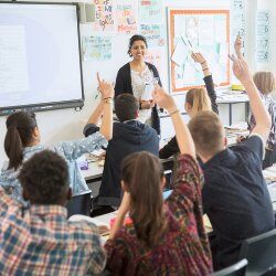 A teacher stands at the front of a class of teenage students who are raising their hands