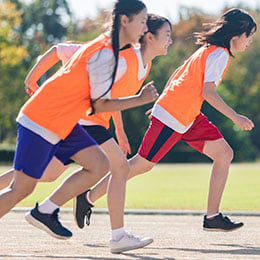 Three young female students running on a track outside wearing orange bibs.