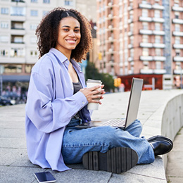 A young woman sits smiling with a laptop on her knee, in front of a blog of apartments